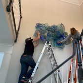 A man and woman hanging an art sculpture in a stairwell